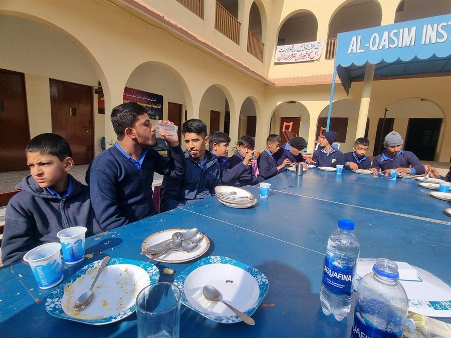 Students seated along a long blue table eating lunch in the courtyard