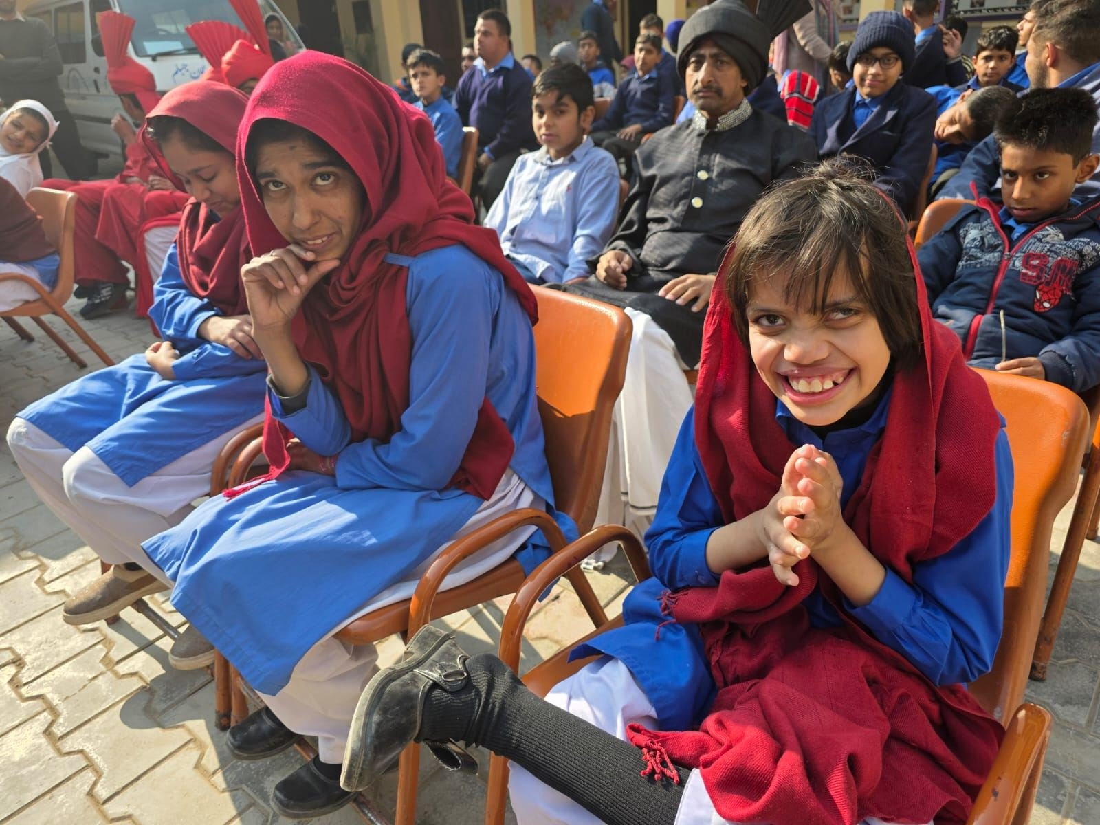 Two students in red shawls seated on orange chairs at an outdoor gathering, one smiling warmly with her hands pressed together